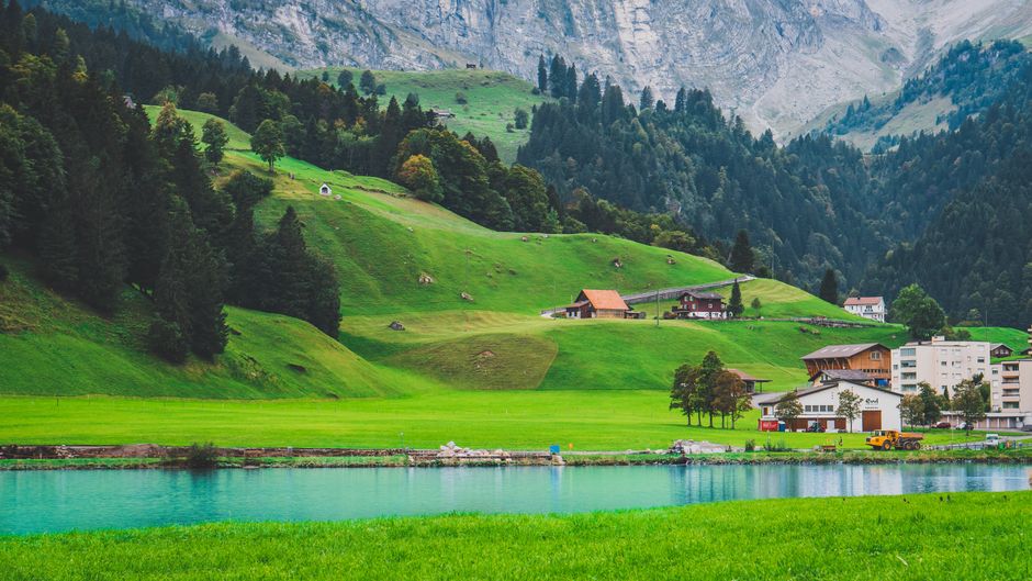 A serene view of lush green hills, a clear blue lake, and quaint houses set against majestic mountains.