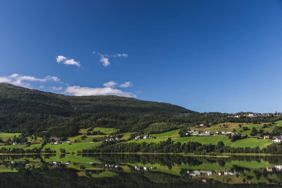Idyllic rural landscape with hills, houses, and lake reflection under a summer sky.